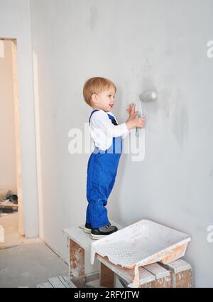 Ein lächelnder kleiner Bauarbeiter streichelt die Wand in einem Apartment, das gerade renoviert wird. Ein bezauberndes Kind im Arbeitsanzug mit Malerwalze bei Hausrenovierungen. Stockfoto