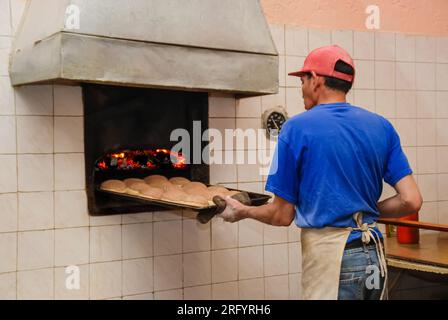 In einer traditionellen mexikanischen Bäckerei dreht sich der Bäcker um, während er frisch gebackene Schokoladen-conchas aus dem Ofen holt. Sein Gesicht bleibt unsichtbar, Emph Stockfoto