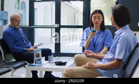 Krankenschwester prüft Blutprobe bei diabetischem asiatischen Mann während des Kontrollbesuchs im Krankenhausbereich. Diabetesrutinkontrolle mit dem Glukometer-Instrument, Assistent bei Blutzuckerspiegelmessung. Stockfoto