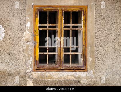 Minimalistische Aufnahme eines einzelnen ländlichen Fensters auf einer grau strukturierten Wand mit abblätterndem Beton. Einfaches Fenster mit gelbem Rahmen und Mullions Stockfoto