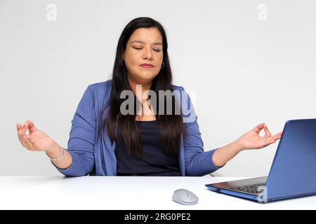 40-jährige Latina-Frau atmet Pranayama ein und übt Yoga in ihrem Büro, um sich zu entspannen und Stress durch Arbeitsdruck abzubauen und Burnout zu verhindern Stockfoto