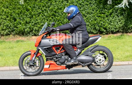 Whittlebury, Northants., Großbritannien - Aug 6. 2023. Ein Mann, der mit einem roten Ducati Motorrad durch ein englisches Dorf fährt. Stockfoto