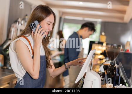 Eine Mitarbeiterin des asiatischen Cafés nimmt eine Vorbestellung per Mobiltelefon entgegen, während sie einen Tablet-Computer in einem Café verwendet. Eine Kellnerin, die am Handy spricht. Lieferkonzept für Heimnahrung Stockfoto