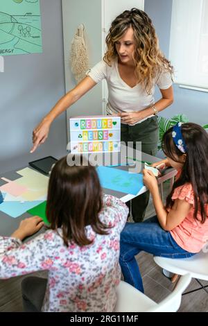 Lehrerin, die den Schülern im Klassenzimmer erklärt, wie es sich um einen ökologischen Prozess handelt, während sie eine Lightbox mit Text Reduce, Reuse, Recycle hält. Nachhaltig l Stockfoto