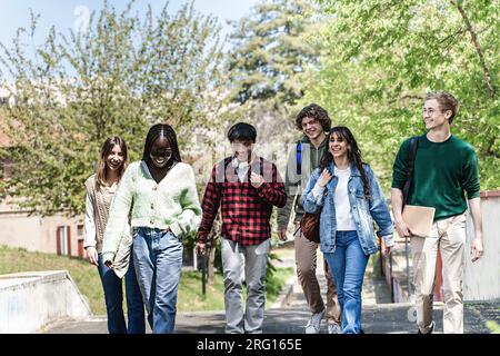 Eine Gruppe von Studenten der Generation Z aus verschiedenen ethnischen Gruppen, mit Rucksäcken auf den Schultern und Notizbüchern in der Hand, durchquert den Park. Stockfoto