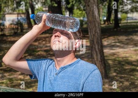 Mann mit Hitze, Typ mit Hitzschlag. Ein weißer Mann presst eine Flasche Wasser ins Gesicht, um sich abzukühlen, er leidet an Hitzewelle, Stuckreiz Stockfoto