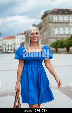 Positive, moderne Seniorin in blauem Kleid, die auf der Straße der Stadt spaziert und hell lächelt, während sie wegblickt Stockfoto