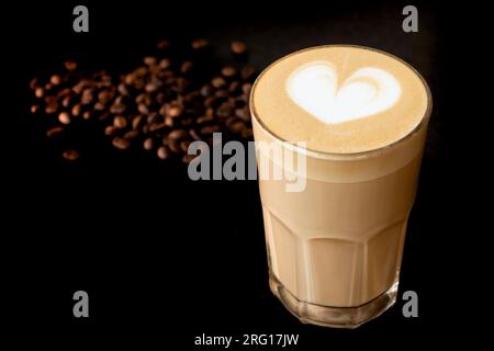 Ein Glas aromatischer, heißer Latte-Kaffee, dekoriert mit Latte Art, auf einem Tisch neben einigen Kaffeebohnen Stockfoto