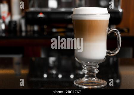Ein Glas aromatischer, heißer Cappuccino auf einem Tisch neben einer Kaffeemaschine auf einem verschwommenen Hintergrund Stockfoto