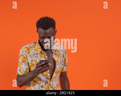 Ein glücklicher junger afroamerikanischer Kerl in einem lässigen Hemd, der sein Smartphone benutzt und auf orangefarbenem Hintergrund steht Stockfoto