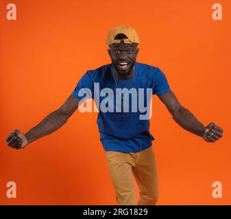 Ein fröhlicher afroamerikanischer Kerl in lässiger Kleidung und Mütze, der vor orangefarbenem Hintergrund in die Kamera schaut Stockfoto