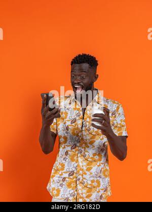 Fröhlicher afroamerikanischer Mann mit Smartphone und Selbstporträt, während er Kartonglas auf orangefarbenem Hintergrund hält Stockfoto