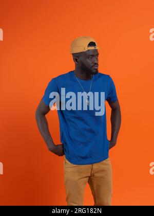Selbstbewusster afroamerikanischer Männchen in lässiger Kleidung, die wegblickt und denkt, während er auf orangefarbenem Hintergrund steht und Hände auf der Taille hat Stockfoto
