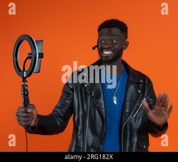 Glücklicher afroamerikanischer Mann in Lederjacke mit Smartphone und LED-Ringlicht mit Stehaufzeichnung auf orangefarbenem Hintergrund einer Studioaufnahme Stockfoto