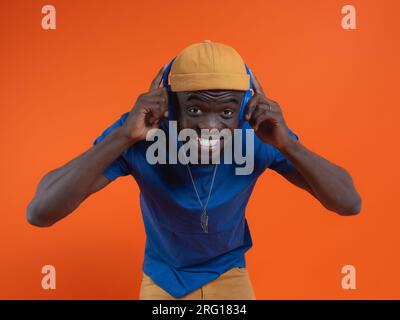 Fröhlicher junger afroamerikanischer Mann in lässigem Outfit und Hut, der fröhlich lächelt, während er auf dem orangefarbenen Hintergrund der Studioaufnahme in die Kamera schaut Stockfoto