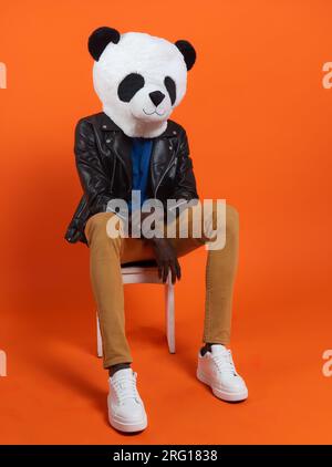 Gesichtslose schwarze Person trägt Panda-Maske, schwarze Lederjacke und weiße Turnschuhe, die auf einem Stuhl vor orangefarbenem Hintergrund im Studio sitzen Stockfoto