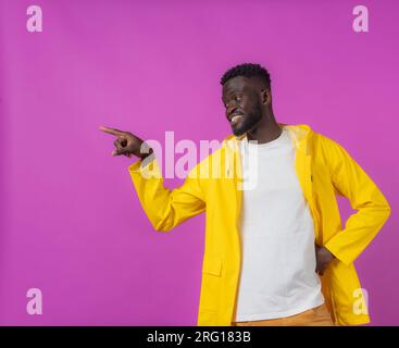 Glücklicher junger afroamerikanischer Mann, der wegblickte, während er mit der Hand auf der Taille stand und mit der Hand auf den lila Hintergrund zeigte Stockfoto