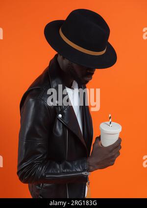 Stylischer, gravierender afroamerikanischer Mann in schwarzer Lederjacke und Hut mit einer Tasse Limonade in der Hand vor einem Studio mit orangefarbenem Hintergrund Stockfoto