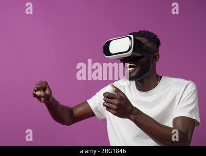 Aufgeregter junger afroamerikanischer Mann in lässiger Kleidung und Virtual-Reality-Headset, während er im Stehen mit der Hand den Controller benutzt und Interes beobachtet Stockfoto