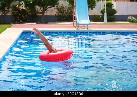 Seitlicher Blick auf eine anonyme Person in rotem Floater, die an sonnigen Tagen in den Swimmingpool mit klarem, blauem, welligem Wasser und auf die Terrasse mit blumigen Bäumen taucht Stockfoto