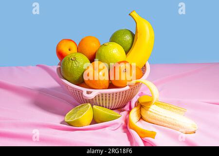 Plastikschüssel mit frischen, sortierten Früchten auf einem Tisch mit rosafarbener Tischdecke bei Tageslicht und blauem Hintergrund Stockfoto