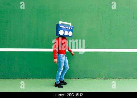 Seitenansicht eines anonymen Mannes in legerer Kleidung, das Gesicht mit blauer Boombox in der Nähe der grünen Wand bedeckt Stockfoto