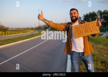 Der Mann trampt am Straßenrand und versucht, das Auto anzuhalten. Er hält Pappe mit Inschrift in der Hand. Stockfoto