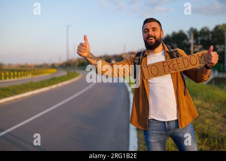 Der Mann trampt am Straßenrand und versucht, das Auto anzuhalten. Er hält Pappe mit Inschrift in der Hand. Stockfoto