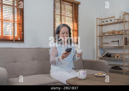 Grauhaarige Asiatin mittleren Alters, die allein auf dem Handy sitzt oder online einkauft, Rentnerin auf dem Sofa im Wohnzimmer Stockfoto