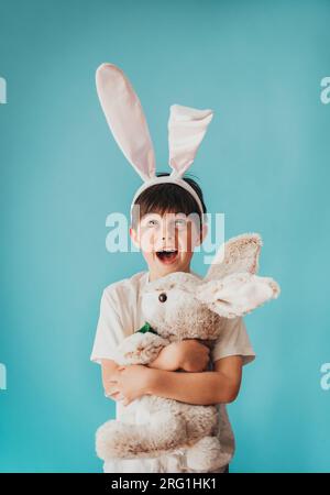 Ein Junge, der ein ausgestopftes Kaninchen hält und auf Hasenohren auf seinem Kopf aufblickt. Stockfoto