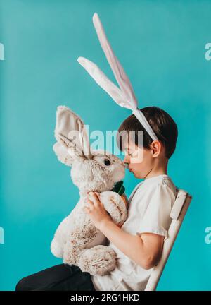 Ein Junge mit Hasenohren, der gefüllte Kaninchen vor blauer Kulisse kuschelt. Stockfoto