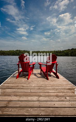 Zwei rote Adirondack-Stühle am Dock mit Blick auf den See an sonnigen Tagen. Stockfoto