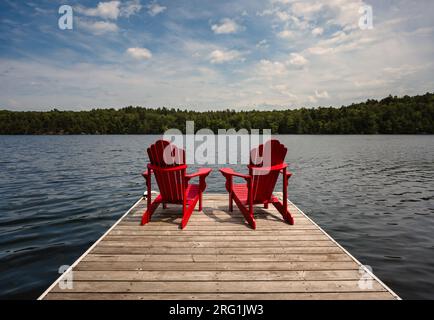 Zwei rote Adirondack-Stühle am Dock mit Blick auf den See an sonnigen Tagen. Stockfoto
