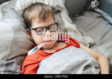 Ein Junge mit Brille liegt auf einem Bett mit einem Thermometer im Mund. Temperaturmessung während einer Kälte Stockfoto