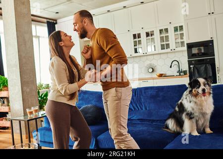 Seitenansicht eines fröhlichen Paares, das sich an der Hand hält und in der Nähe des Border Collie Hundes auf dem Sofa zu Hause tanzt Stockfoto