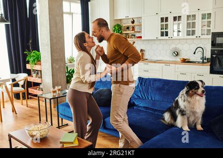 Lächelndes Paar, das Händchen hält und in der Nähe des Border Collie auf dem Sofa im Wohnzimmer zu Hause tanzt Stockfoto