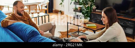 Lächelnde Frau streichelt Border Collie Dog auf der Couch neben Freund im Wohnzimmer, Banner Stockfoto