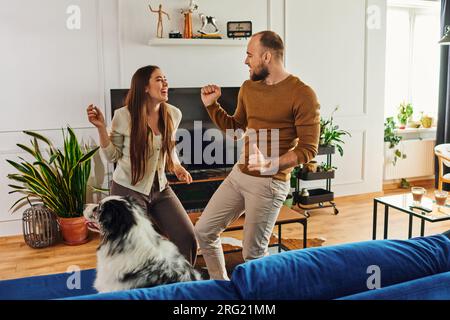 Fröhliches Paar in lässiger Kleidung, das in der Nähe von Border Collie auf dem Sofa im Wohnzimmer zu Hause tanzt Stockfoto