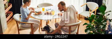 Lächelnder Mann in Haushaltskleidung, streichelnder Border Collie, während er mit seiner Freundin zu Hause frühstückt, Banner Stockfoto
