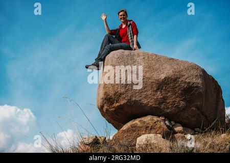 Eine junge Touristin sitzt auf einem großen Felsbrocken und winkt mit der Hand Stockfoto