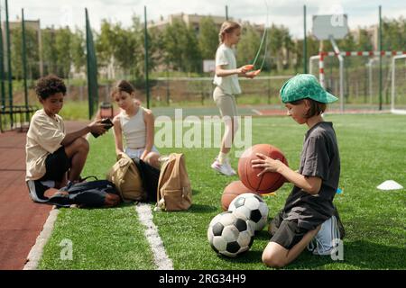 Seitenansicht eines Schuljungen in Sportbekleidung, der den Ball für das Sportspiel auswählt, während er auf dem grünen Rasen des Fußballfelds auf den Knien steht Stockfoto