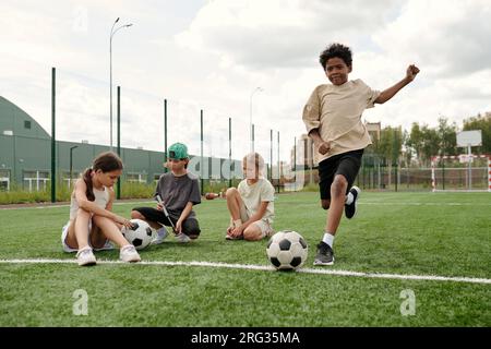Afroamerikanischer Schuljunge in Aktivbekleidung, der nach dem Fußball auf dem grünen Fußballfeld läuft, während er gegen seine Freunde ein Sportspiel spielt Stockfoto