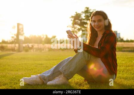 Eine Frau, die an einem Sommertag auf grünem Gras im Park sitzt und bei Sonnenuntergang Musik hört. Stockfoto