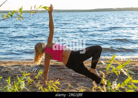 Eine Rückansicht einer Frau, die auf ihren Händen steht und ihren Arm hochstreckt. Eine Frau, die im Sommer am See Yoga praktiziert. Meditation. Sommersport Stockfoto