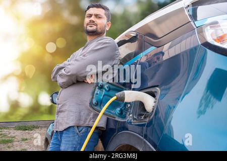 Ein Indianer steht, während sein modernes Elektroauto an der Ladestation aufgeladen wird. Ladeanschluss für die Batterie des Elektrofahrzeugs. Erneuerbares Olen Stockfoto