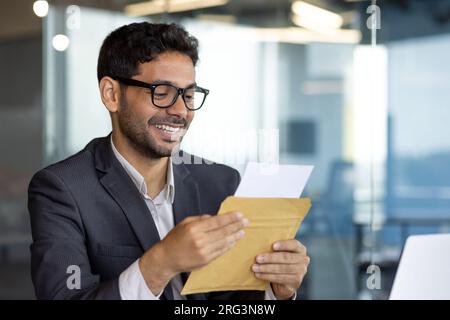 Junger erfolgreicher arabischer Geschäftsmann am Arbeitsplatz im Büro, ein lächelnder Mann, der mit Zufriedenheit eine Nachricht mit einer Mail-Benachrichtigung mit einer guten Nachricht erhalten hat, Chef im Business-Kostüm. Stockfoto