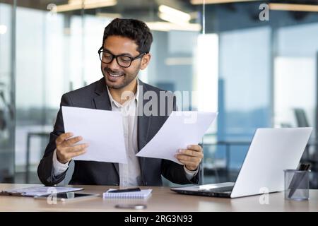 Erfolgreicher, erfahrener Finanzier in Papierarbeit, arabisches Lächeln, zufrieden mit den Ergebnissen der finanziellen Errungenschaften, Prüfung von Verträgen, Rechnungen und Kontoauszügen, Geschäftsmann in Anzug. Stockfoto