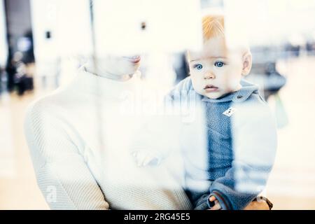 Die nachdenkliche junge Mutter schaute durch das Fenster und hielt sein kleines Kind fest, während sie darauf wartete, an den Abflugsteigen des Flughafenterminals an Bord eines Flugzeugs zu gehen. Das „Reise mit Baby“-Konzept Stockfoto