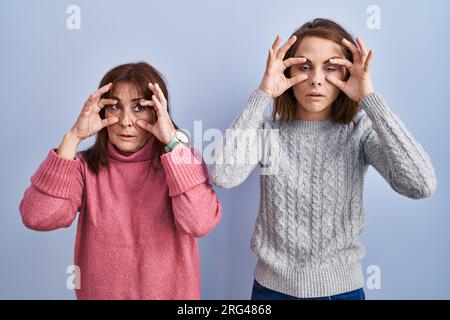 Mutter und Tochter stehen auf blauem Hintergrund und versuchen, die Augen mit den Fingern zu öffnen, schläfrig und müde wegen Morgenmüdigkeit Stockfoto