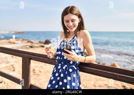 Junge blonde Touristin, die Eis am Meer mit einem Smartphone hält Stockfoto